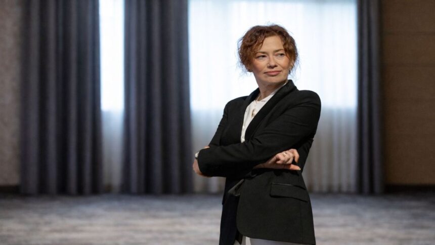 Portret editorial al Irinei Vincze, îmbrăcată într-un costum negru, surprinsă într-o postură calmă și sigură, cu brațele încrucișate, într-un spațiu interior sobru, sugerând reflecție și autoritate intelectuală.