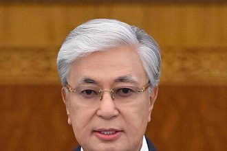 President Kassym-Jomart Tokayev delivering a formal address, wearing a dark suit and blue tie, with Kazakhstan flag pin on his lapel.