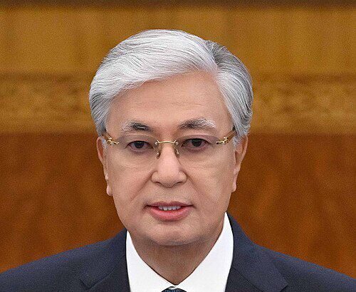 President Kassym-Jomart Tokayev delivering a formal address, wearing a dark suit and blue tie, with Kazakhstan flag pin on his lapel.