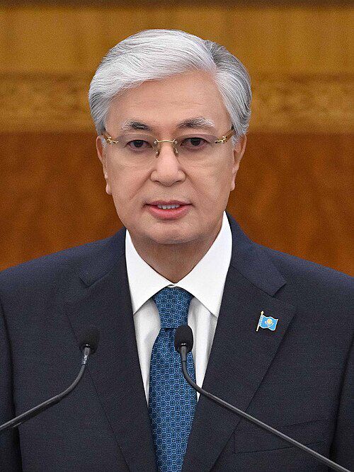 President Kassym-Jomart Tokayev delivering a formal address, wearing a dark suit and blue tie, with Kazakhstan flag pin on his lapel.